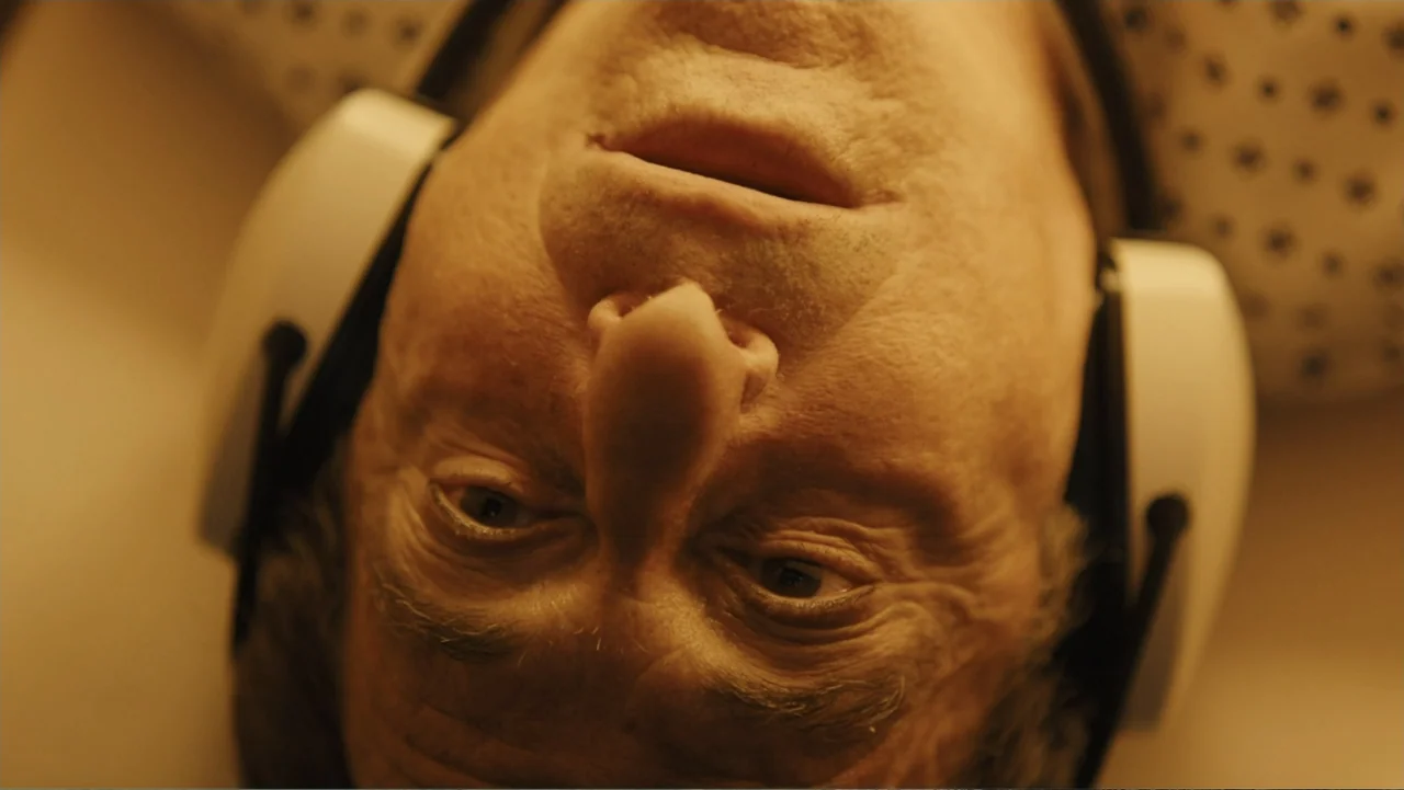 Paterno (2018) movie still: closeup — An interior space with a person lying on their back. An older man wearing large white headphones looks…