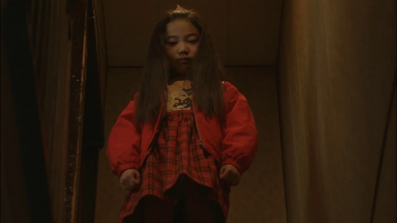 Battle Royale (2000) movie still: medium — A dimly lit narrow stairwell with textured brown walls and low light levels. A young girl stands on the…