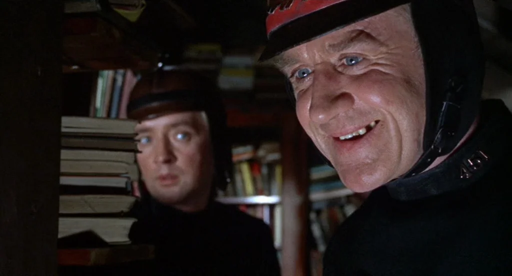 Fahrenheit 451 (1966) movie still: medium — A dimly lit room features floor-to-ceiling wooden bookshelves packed with rows of old books. Two men…
