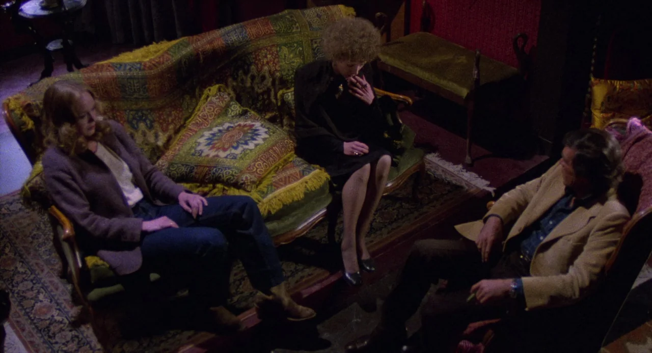 City of the Living Dead (1980) movie still: A group of three people sit in a cluttered, dim, red-hued room, caught in an moment of heavy, unspoken…