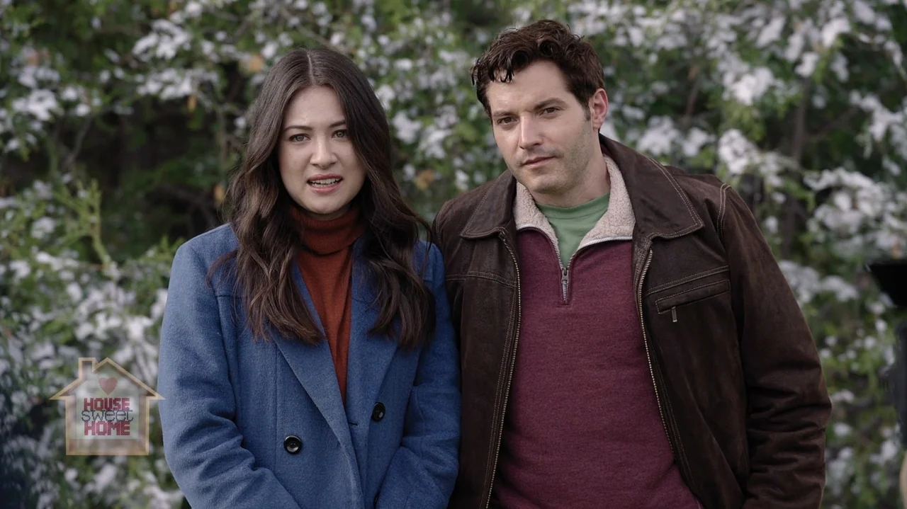 Designing Christmas (2022) movie still: medium — A man and a woman stand outdoors in front of a blooming white-flowering bush. The two individuals…