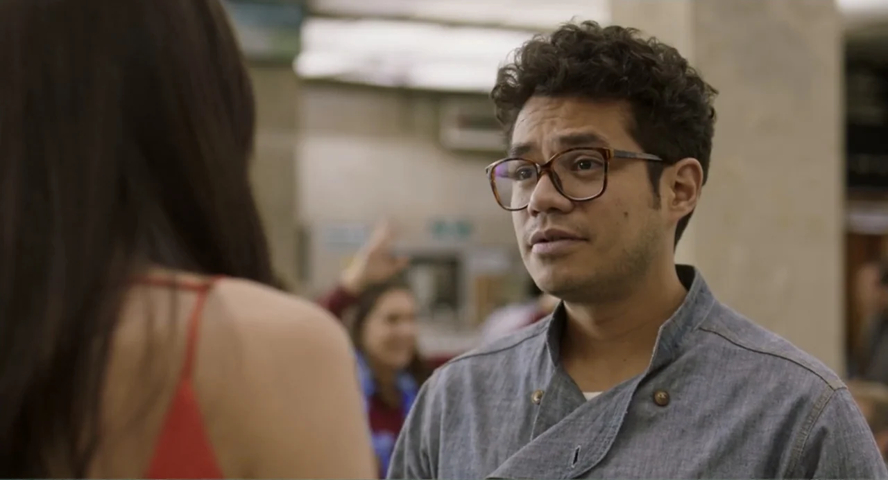 Casando a mi Ex (2023) movie still: medium — A man stands in a brightly lit indoor public area with other people blurred background. The man in a…