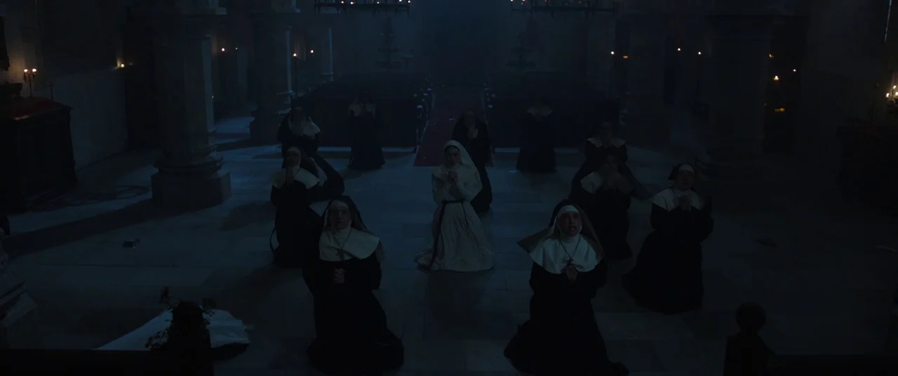 The Nun (2018) movie still: wide — A dark, expansive stone cathedral nave features rows of kneeling figures in religious garments. Twelve nuns pray…