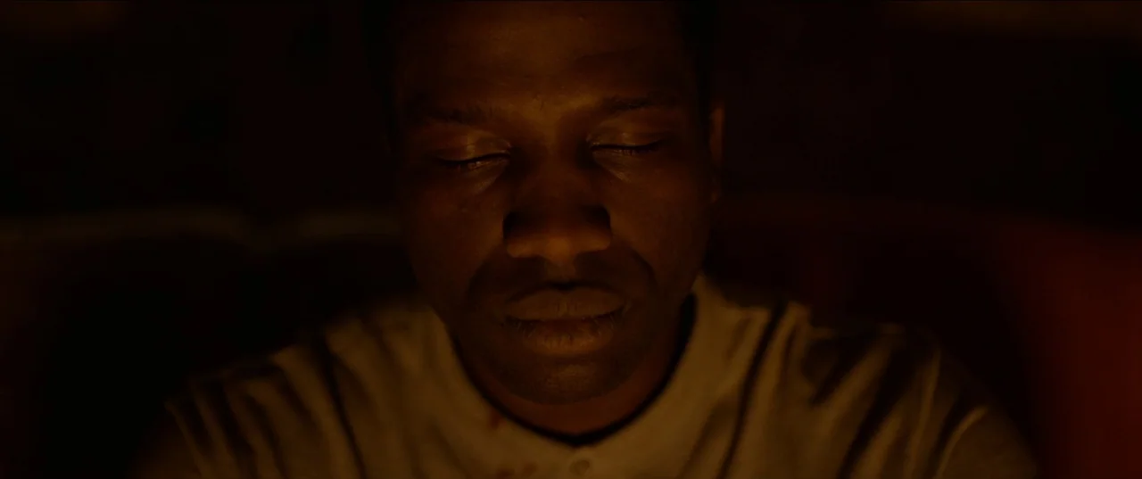 His House (2020) movie still: closeup — A dimly lit interior space contains a man centered in the frame. The man has his eyes closed and faces the…