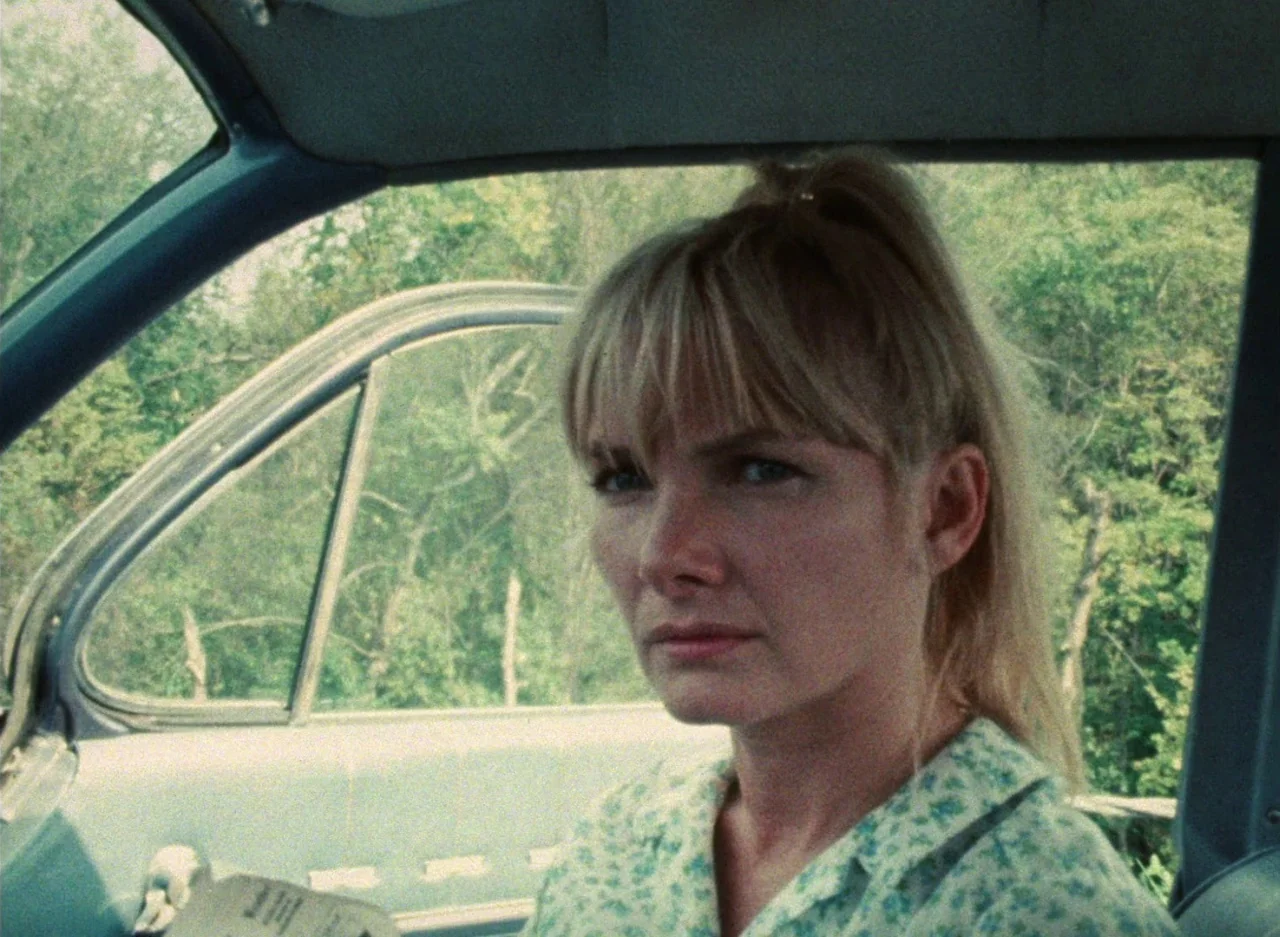 Wanda (1970) movie still: medium — The interior of a 1970s sedan features a view of lush green trees outside. A young woman sits in the driver's…