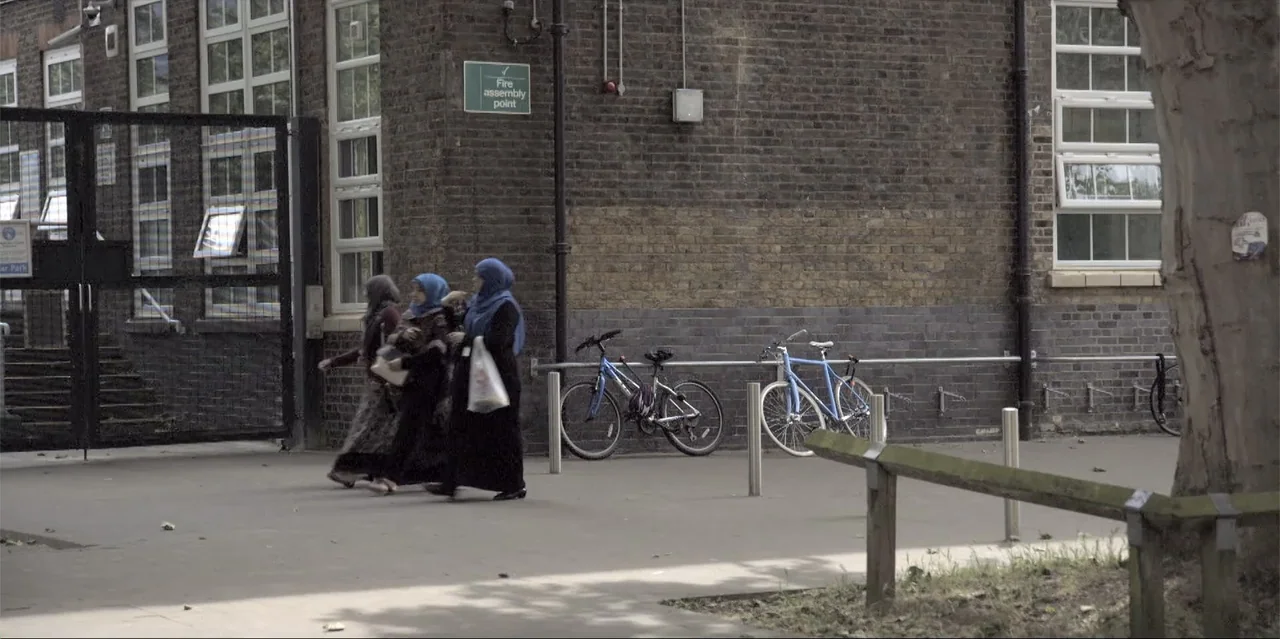 A Radical Life (2022) movie still: fullbody — A brick building wall features a green sign and bicycle racks in the background. Three women in long…