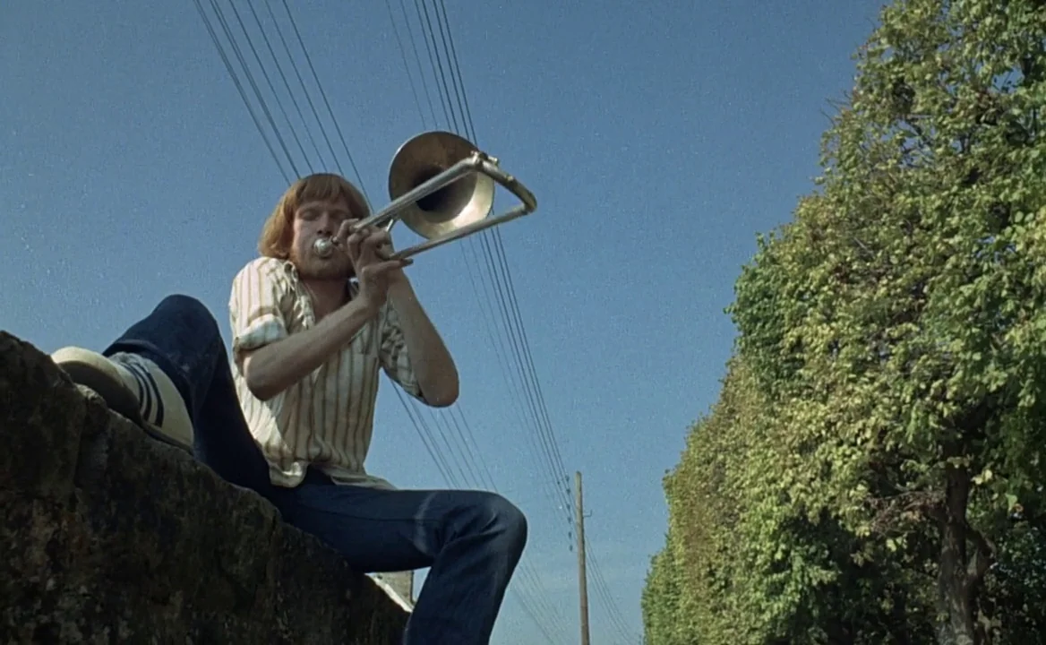 F for Fake (1973) movie still: medium — A man sits on a stone wall next to a line of trees under blue sky. He holds and plays a metal trombone with…