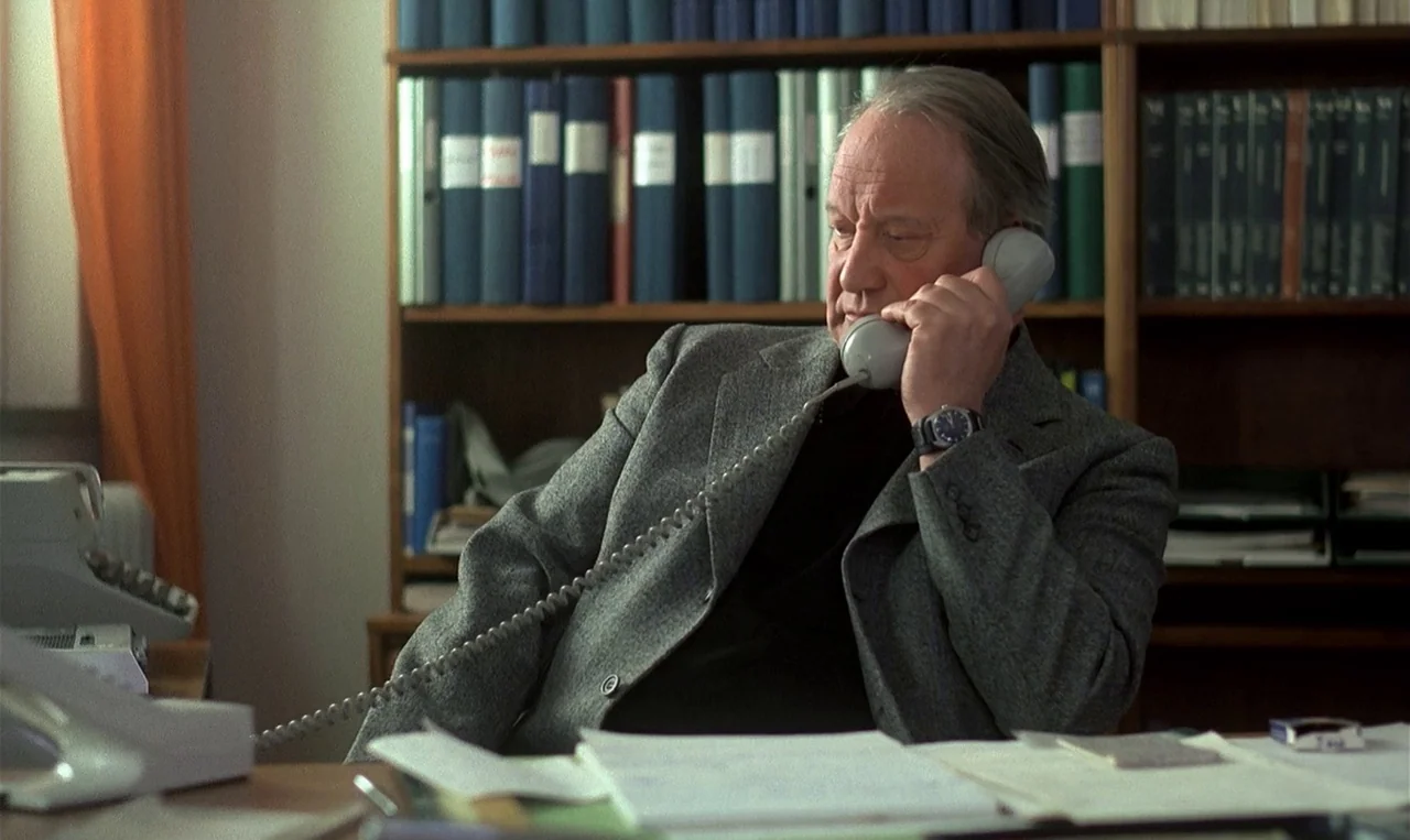 Man on the Roof (1976) movie still: medium — A cluttered desk sits before a wooden bookshelf filled with many blue-bound binders. An older man in a…