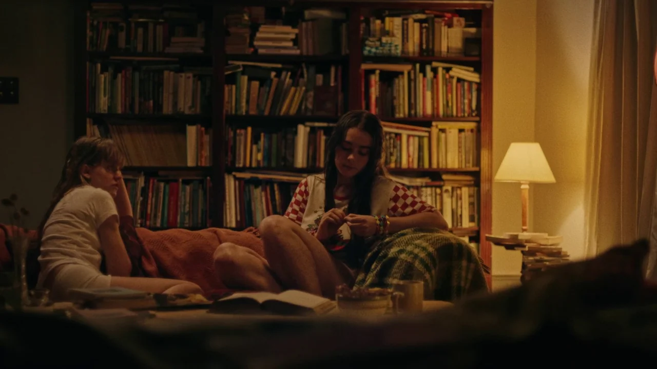 My First Summer (2020) movie still: medium — A domestic space features a large, fully stocked floor-to-ceiling wooden bookshelf. Two young women sit…