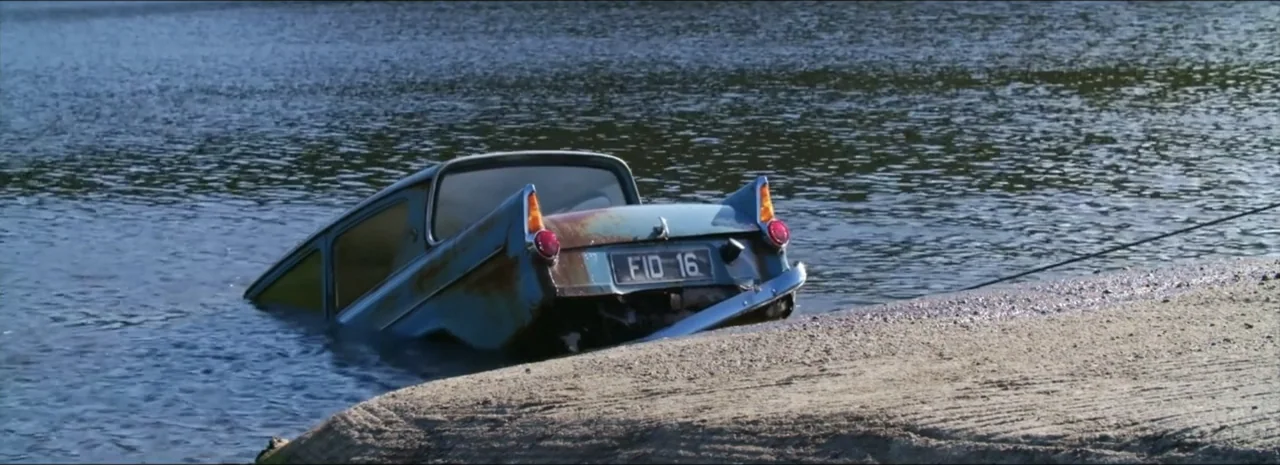 DannyBoy (2020) movie still: wide — A light blue vintage sedan rests partially submerged in water against a sandy shoreline. A thin cable extends…