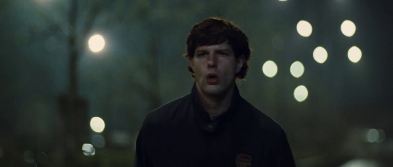 Our Day Will Come (2010) movie still: medium — A dark outdoor space at night with blurred streetlights in the background. A young man runs through…