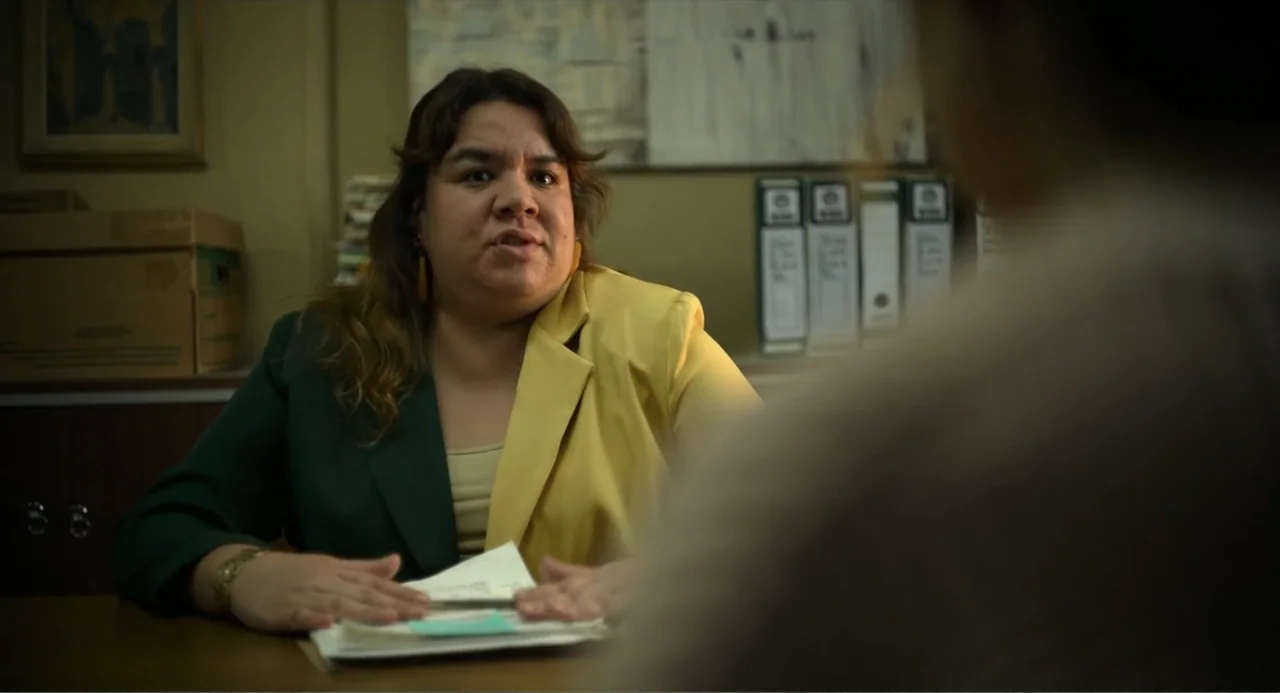Heartbeat (2008) movie still: medium — A dimly lit office features a desk with stacks of documents and filing folders behind. A woman sits behind…