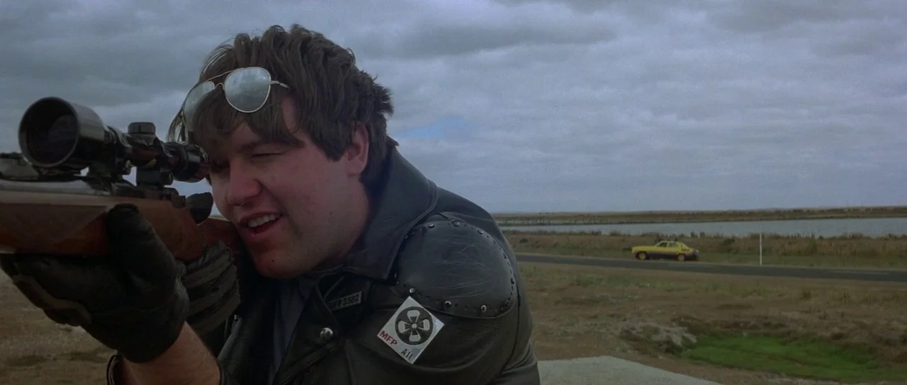Mad Max (1979) movie still: medium — An overcast, flat landscape stretches out along a paved road near water. A man in a leather jacket kneels while…