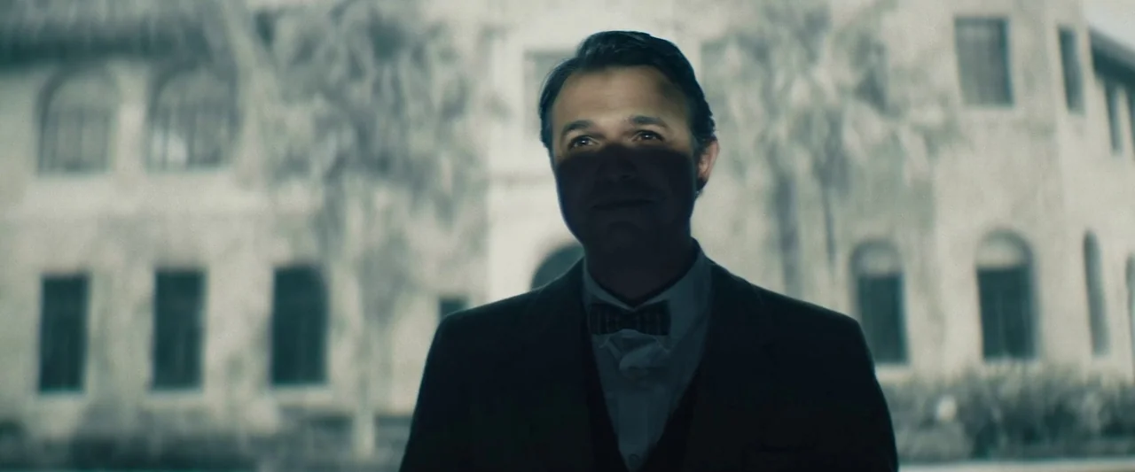 The Mad Hatter (2021) movie still: medium — A man stands before a large, multi-story masonry building with numerous arched windows. The subject…