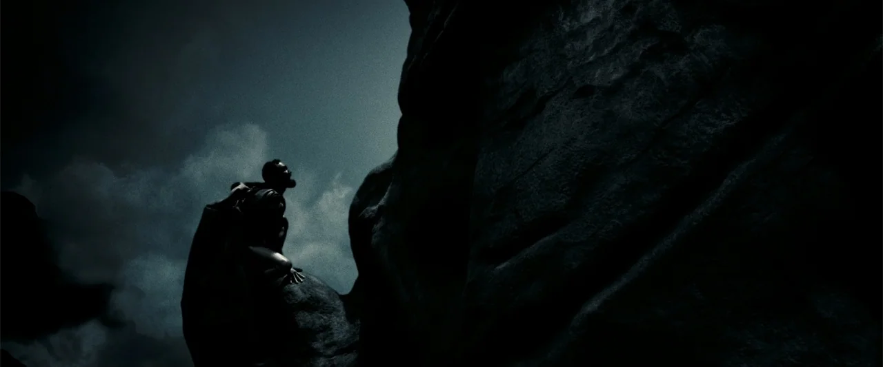 300 (2007) movie still: wide — A dark rocky landscape features a towering cliff face against a cloudy sky. A solitary man crouches on a rock outcrop…