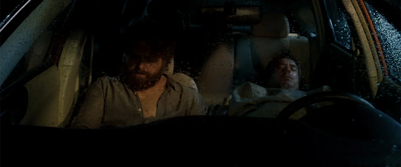 Due Date (2010) movie still: medium — The interior of a vehicle at night with rain droplets visible on the windshield. Two men are asleep in the…