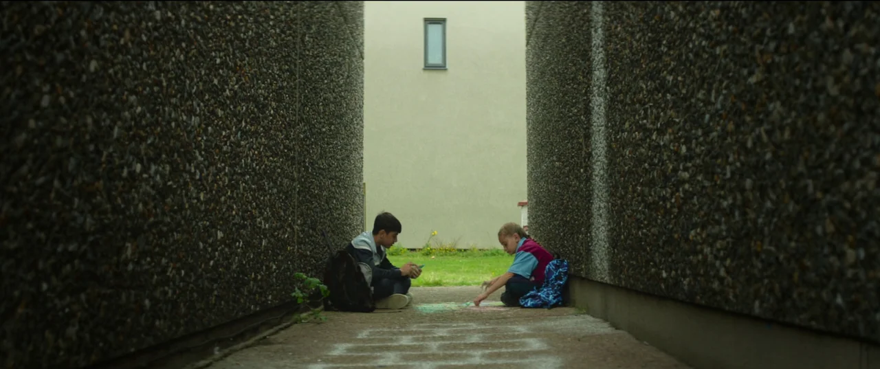 Scrapper (2023) movie still: wide — A narrow outdoor concrete passageway is flanked by high, rough-textured grey aggregate walls. Two children sit…