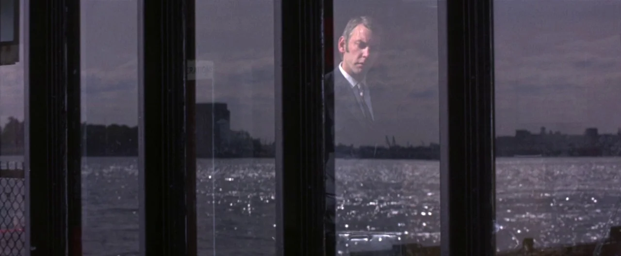 Klute (1971) movie still: medium — A man stands inside a building behind large glass windows overlooking a body of water. The reflection of the…