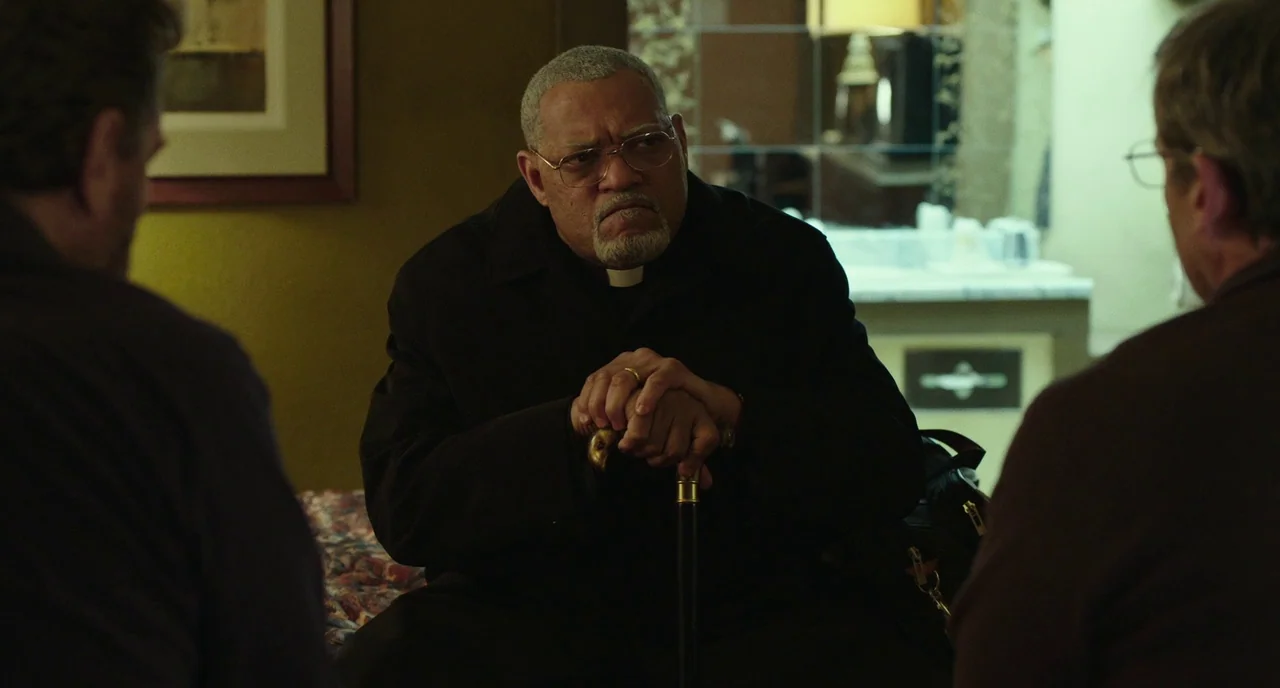 Last Flag Flying (2017) movie still: medium — A dark motel room with a bed and a blurred background counter area. An older priest wearing a clerical…