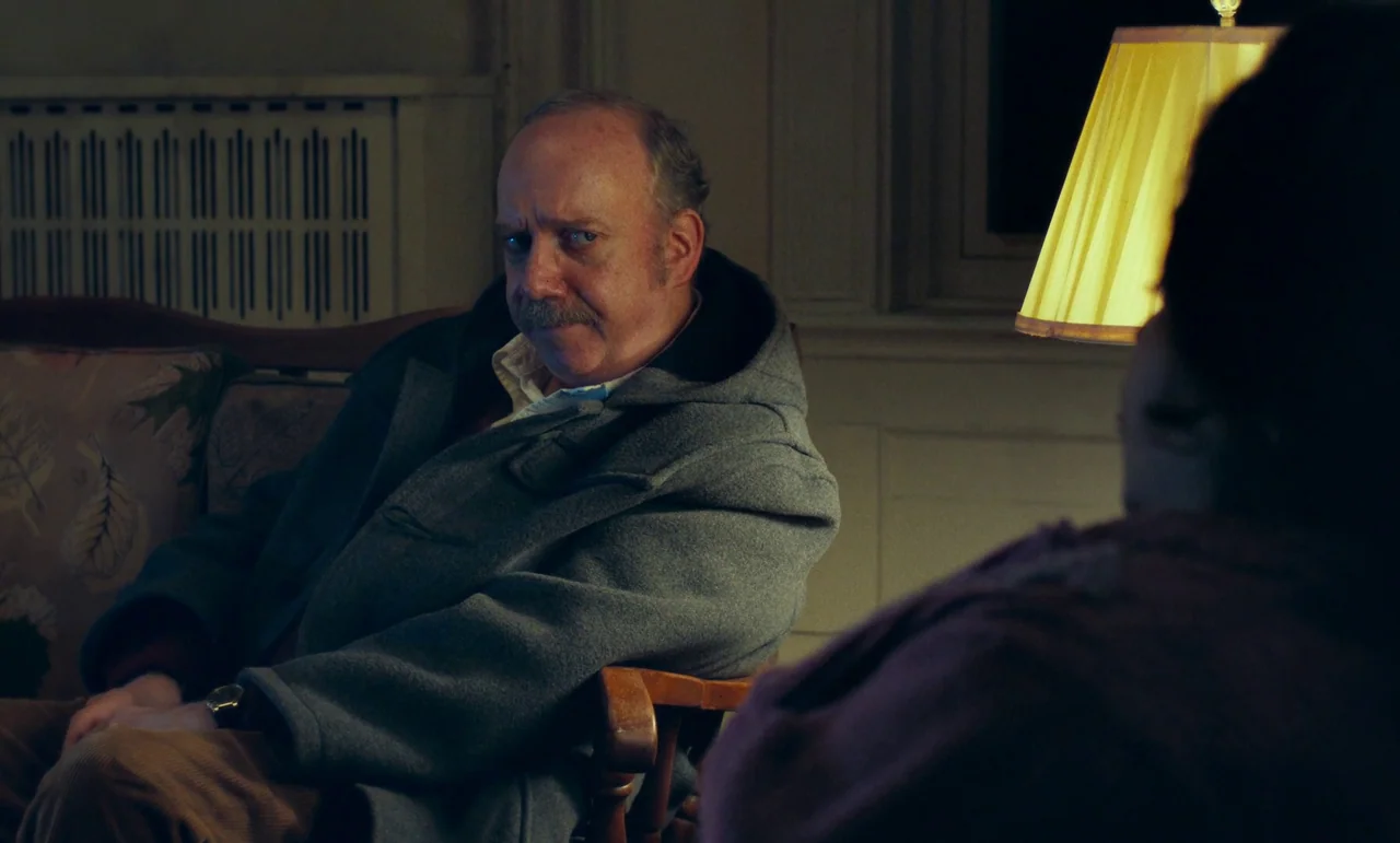 The Holdovers (2023) movie still: medium — A dimly lit, wood-paneled room is illuminated by a yellow-shaded lamp. A middle-aged man in a grey wool…