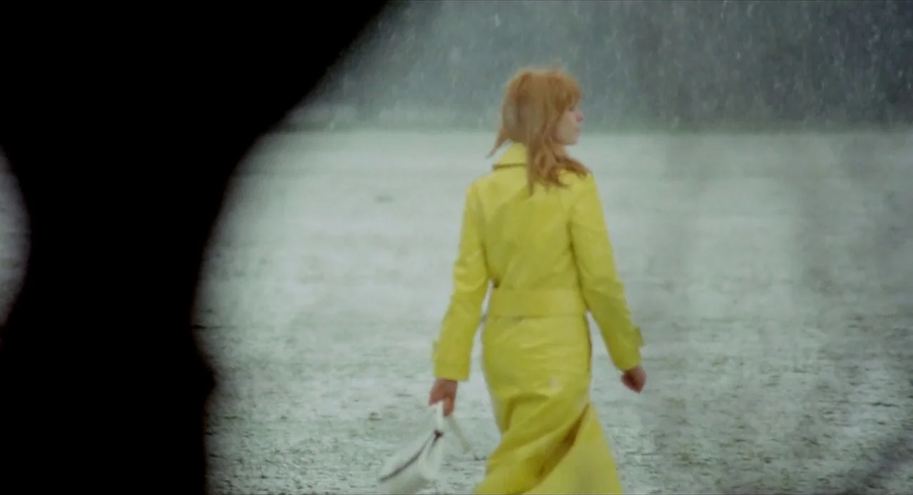 Deep End (1970) movie still: fullbody — A woman in a bright yellow raincoat walks across an open grassy field. Rain falls steadily while a blurred…