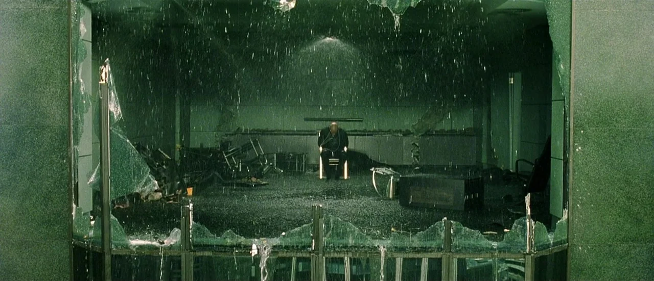 The Matrix (1999) movie still: wide — A dark office space features shattered floor-to-ceiling glass panels and heavy interior rain flooding. A…