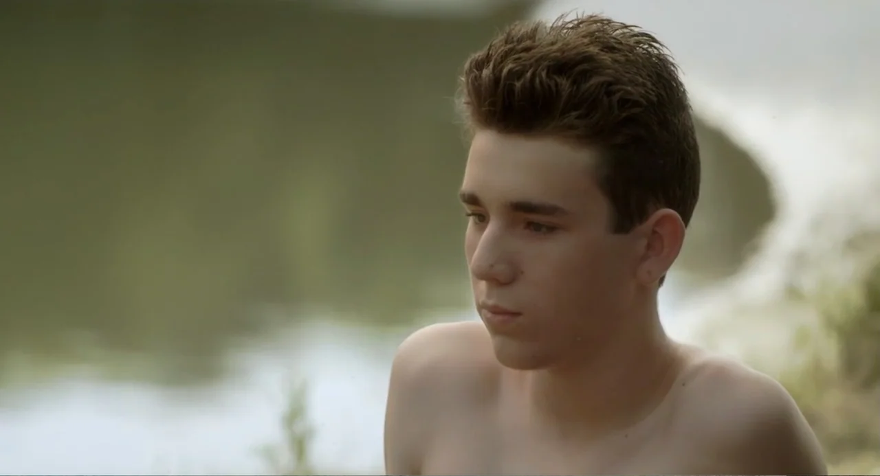 Newton's Third Law (2017) movie still: medium — A lakeside bank features blurred natural greenery and water in the background. A shirtless teenager…