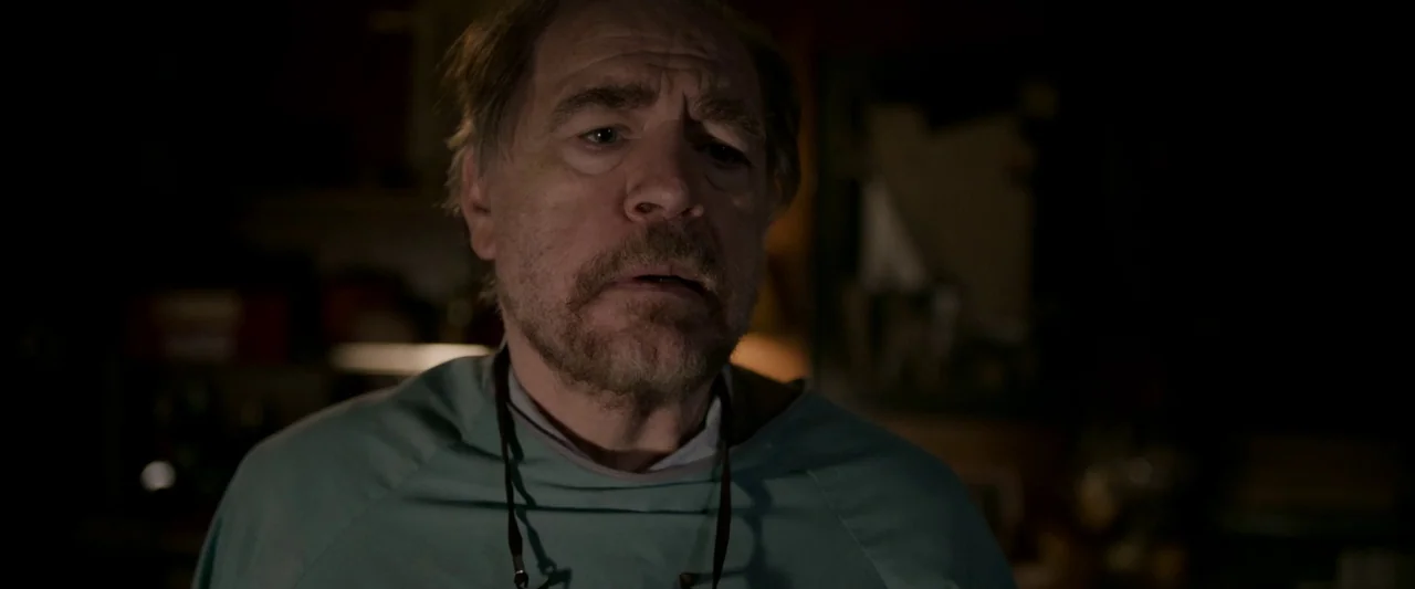 The Autopsy of Jane Doe (2016) movie still: closeup — A dimly lit interior space with an obscured, dark background and neutral tones. An older man…