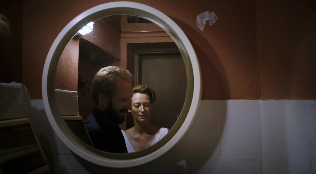 A Bigger Splash (2015) movie still: medium — A small bathroom with a plain white tiled wall and dark brown painted upper section. A man with a beard…