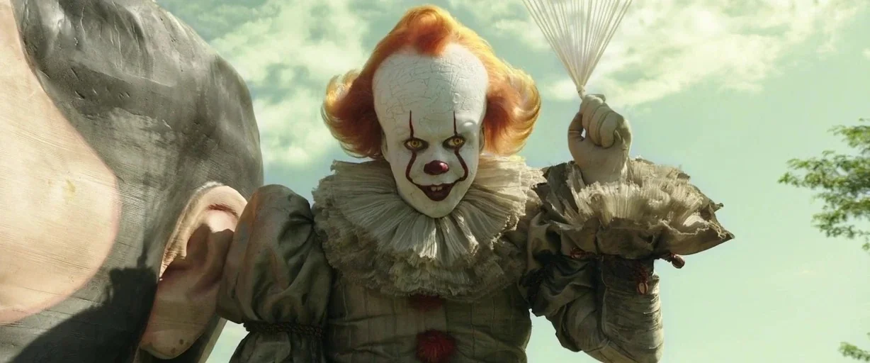 It Chapter Two (2019) movie still: medium — An outdoor location features a clown standing next to a large, unidentifiable object. The clown holds a…