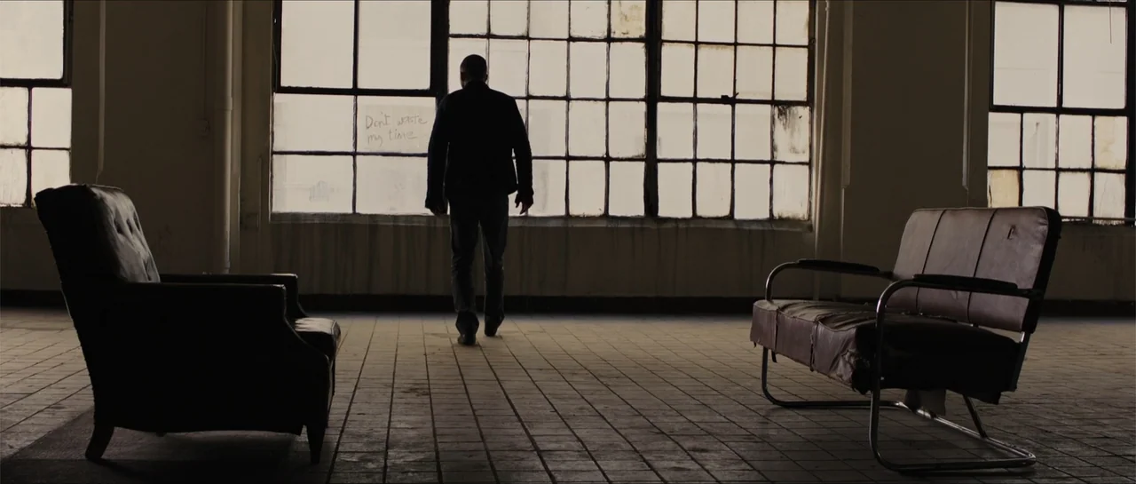In Time (2011) movie still: fullbody — An abandoned industrial space has grid windows and tiled floors with worn furniture. A solitary man walks…