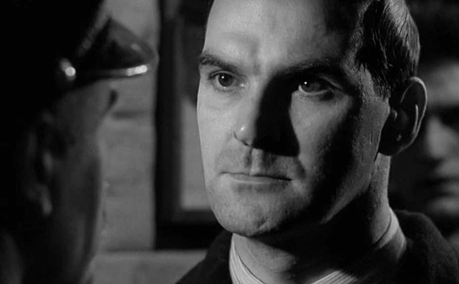 The Criminal (1960) movie still: closeup — A dimly lit room with high-contrast shadows obscuring parts of the subjects. A man faces another…