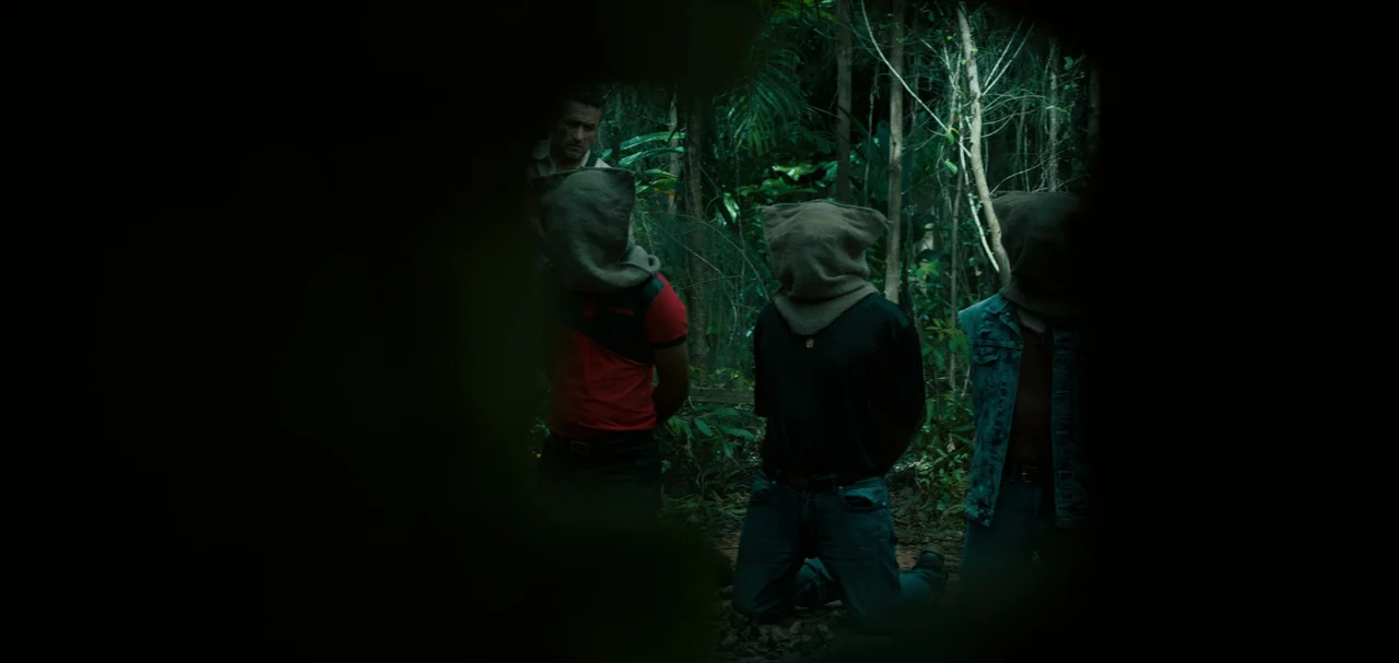 Triple Frontier (2019) movie still: medium — Three individuals kneel on the jungle floor with burlap bags covering their heads. An armed man watches…