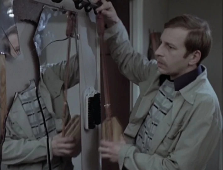 Camera Buff (1979) movie still: medium — An indoor space features a wall-mounted mirror partially broken and cracked at the edges. A man in a tan…