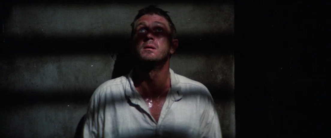 Papillon (1973) movie still: medium — A dimly lit stone enclosure features horizontal bars casting shadows across the interior wall. A man in a…