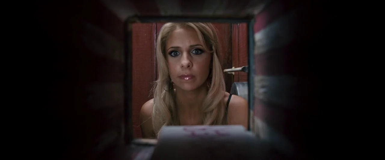 Southland Tales (2006) movie still: closeup — A woman looks through a rectangular opening in a wooden structure. The wooden frame is dark and narrow…