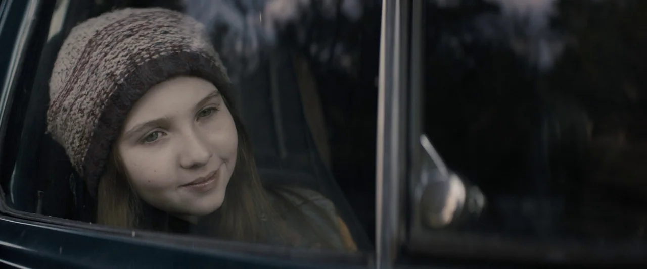 Don’t Breathe 2 (2021) movie still: closeup — A view from outside a vehicle into the passenger cabin. The girl leans against the window frame while…