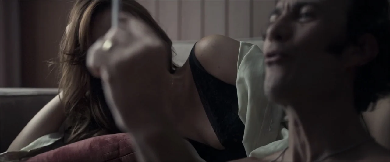 Angela (2023) movie still: closeup — A dark bedroom interior features a man and woman lying on a bed. The couple is positioned close together while…