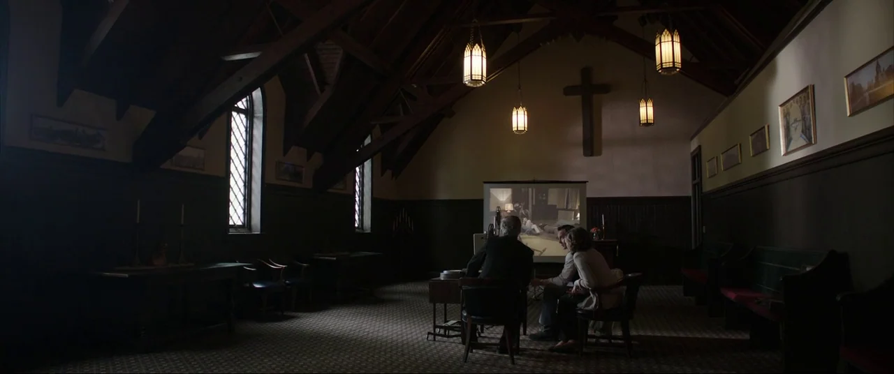 The Conjuring (2013) movie still: wide — A quiet, dark parish hall features a wooden cross on the wall and wooden beams. Three people sit together…
