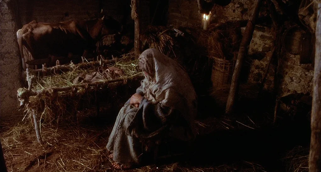 Life of Brian (1979) movie still: medium — A dark, rustic stable interior filled with scattered straw and rough wooden supports. A woman draped in…