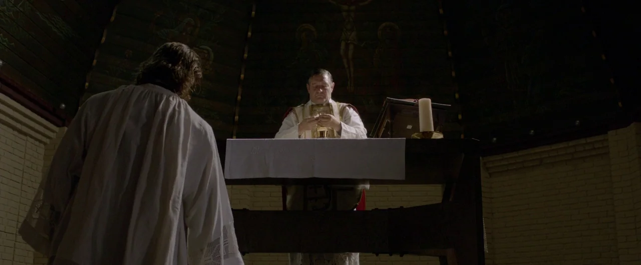 Borgman (2013) movie still: medium — A dark, circular chapel features a wooden altar centered beneath a mural of a crucifixion. A priest stands…