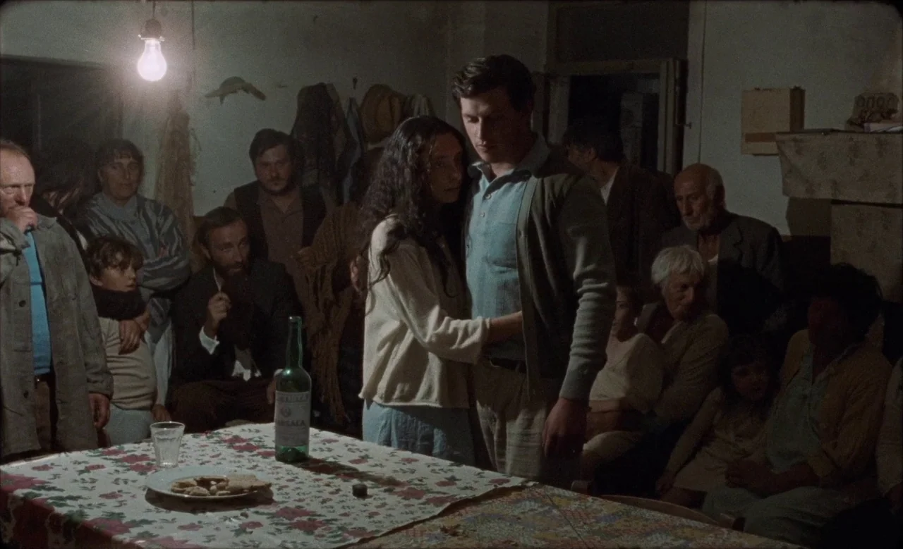 Happy as Lazzaro (2018) movie still: medium — A dimly lit, rustic room features a large group of people gathered around a table. A man and a woman…