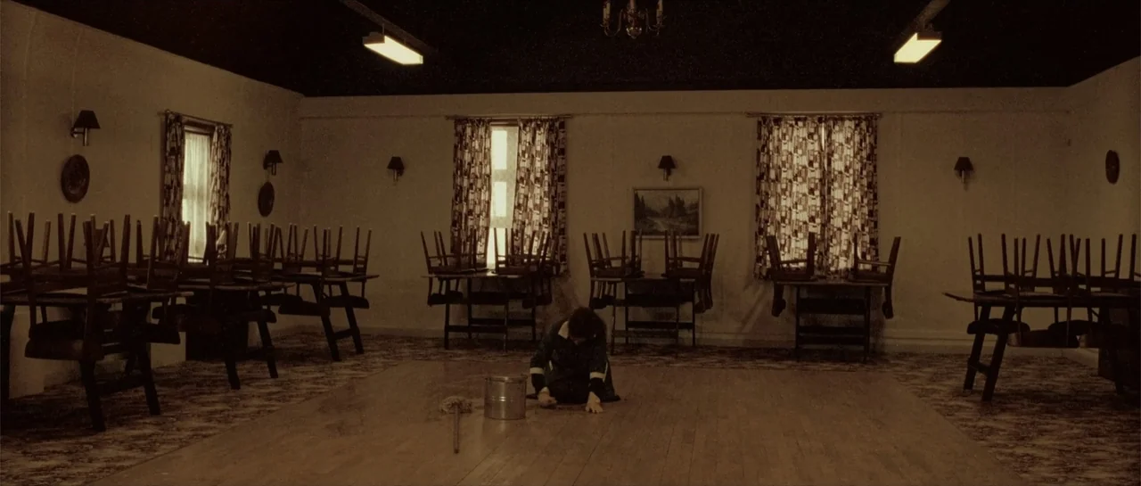 Breaking the Waves (1996) movie still: wide — An empty dance hall features rows of wooden tables with chairs stacked on top. A lone worker kneels on…