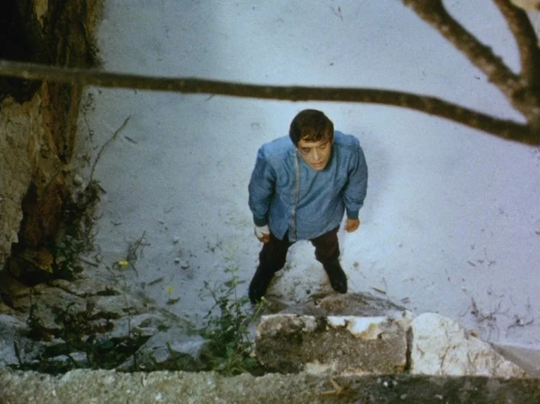 Our Lady of the Turks (1968) movie still: fullbody — A man stands in a sandy, recessed area enclosed by rocky, textured walls. He looks directly…