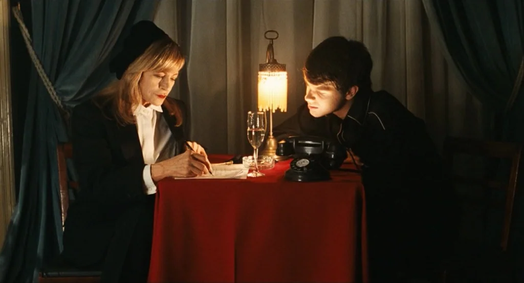 This Night (2008) movie still: medium — A table covered in a red cloth sits between two people in a room. The woman writes a note while the young…
