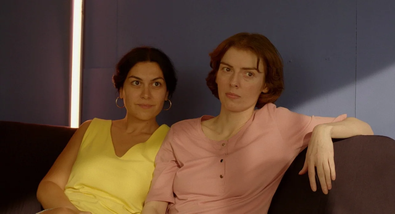 Blackbird Blackbird Blackberry (2023) movie still: medium — Two women sit together on a sofa against a flat blue wall. A bright vertical light…