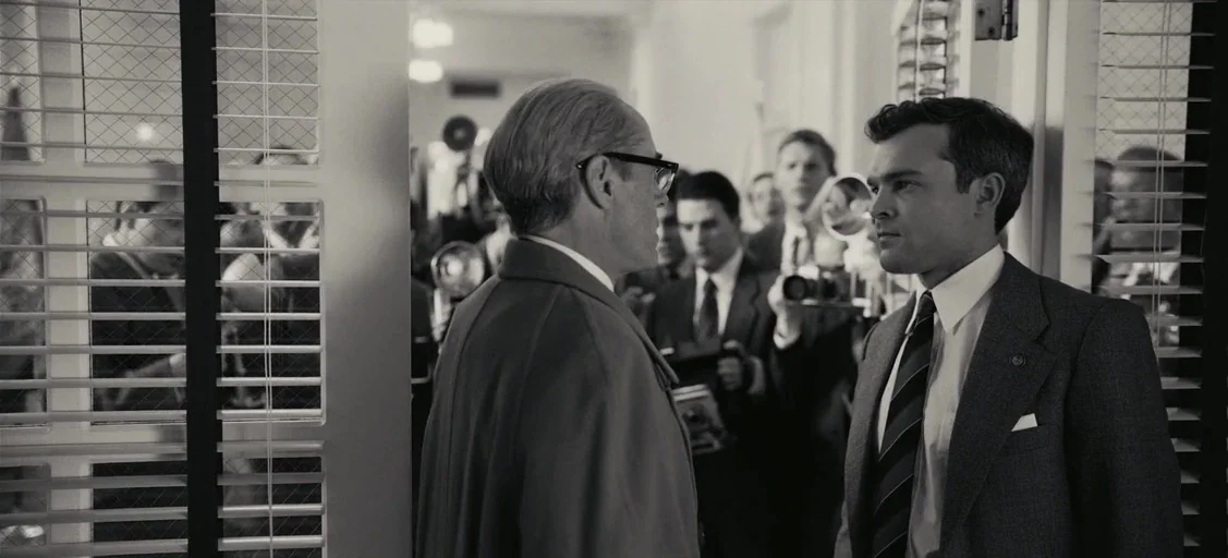 Oppenheimer (2023) movie still: medium — A crowded room with people holding cameras framed through a partially open doorway. Two men stand in the…