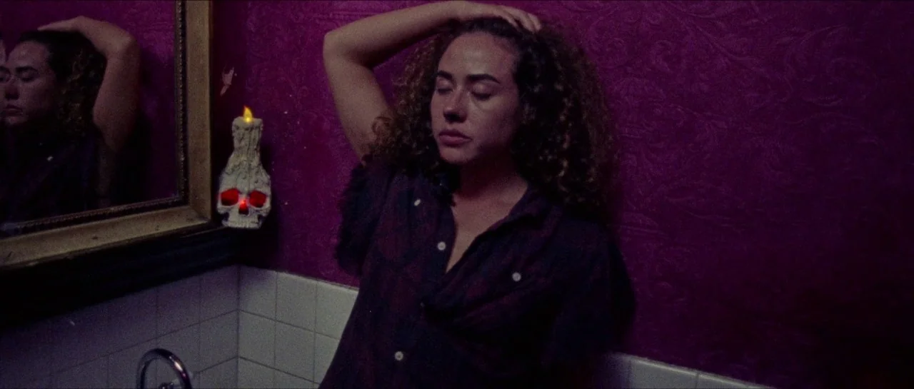 Bliss movie still: medium — A bathroom featuring dark magenta wallpaper with a patterned texture and white tiled counters. A young woman with curly…