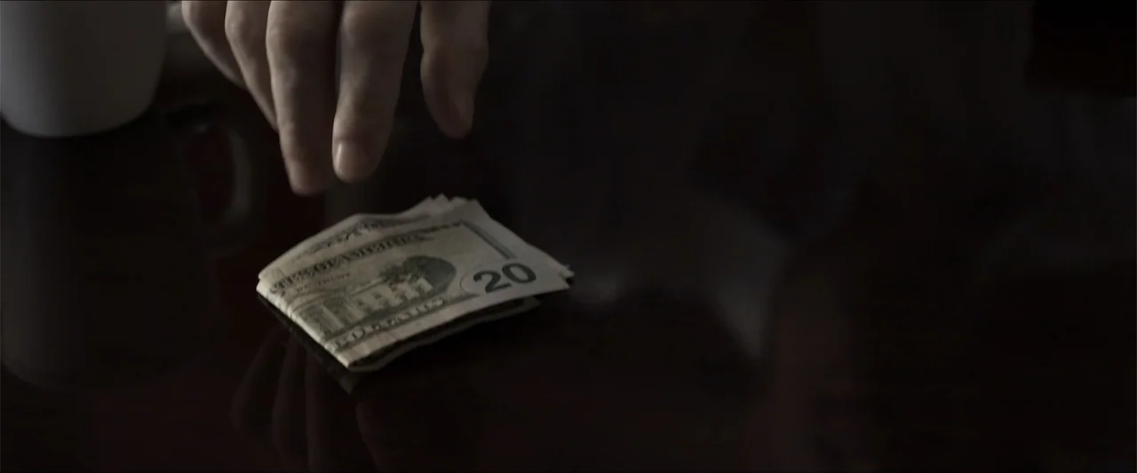 Heavenly Deposit (2019) movie still: closeup — A dark, reflective table surface holds a folded twenty dollar bill. A hand descends toward the money…