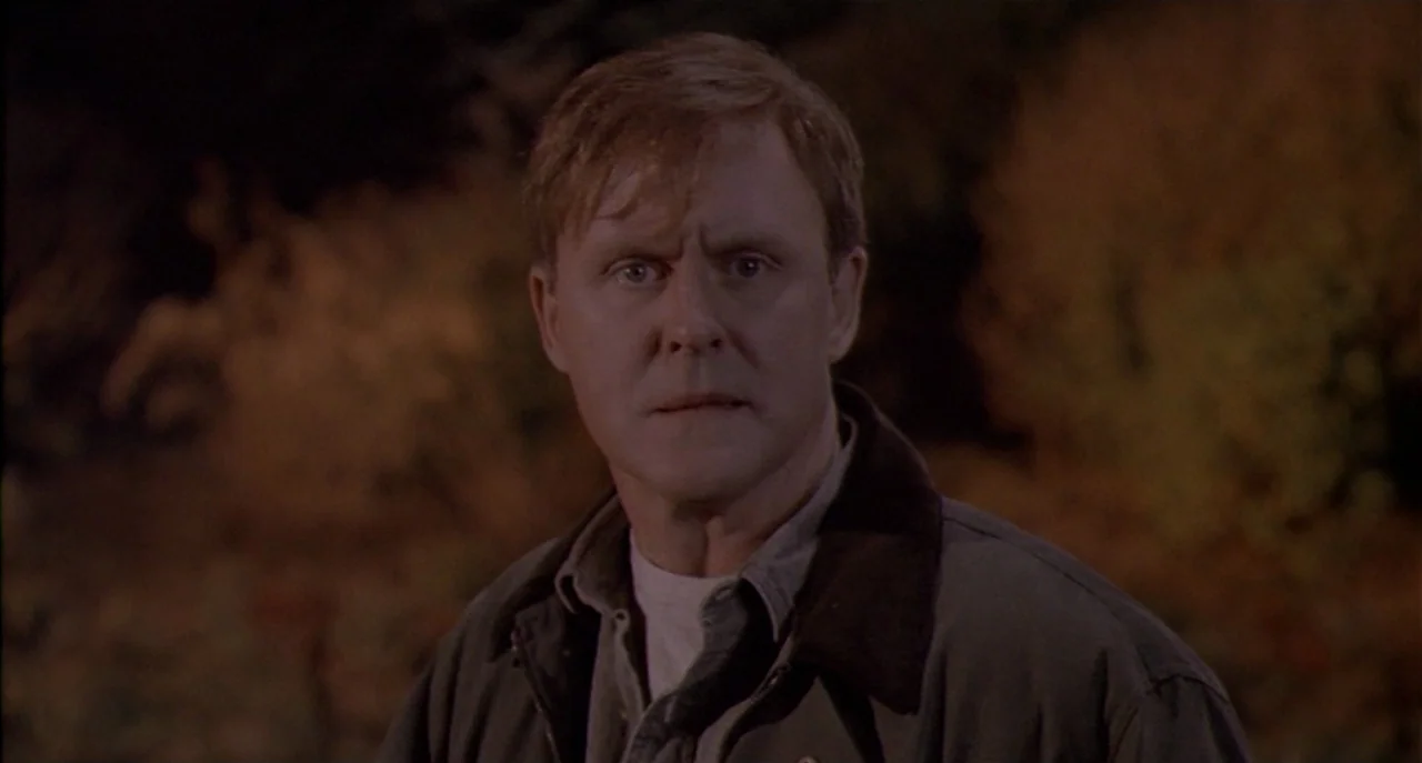 Raising Cain movie still: medium — A dark outdoor setting features a man standing in front of out-of-focus foliage. The man maintains a steady gaze…